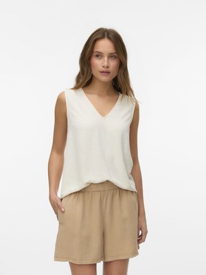 Vrouw staand, gekleed in een mouwloos wit topje met V-hals en beige shorts met een elastische taille, met haar linkerhand in haar zak.