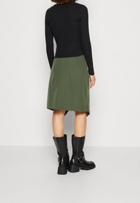Top lungo a maniche ribbed nera abbinato a una gonna verde oliva. La gonna presenta un orlo leggermente asimmetrico. Stivali neri al ginocchio con dettagli hardware.