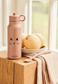 Różowa butelka na wodę ze stali nierdzewnej z wzorem twarzy kota, obok miski z żółtymi gruszkami i różową łyżką na kremowej tkaninie.