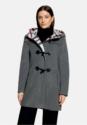 Femme portant un manteau en laine gris avec des attaches à bascule et une capuche doublée d’un tissu à carreaux rouge, blanc et bleu, associé à un pantalon noir et un col roulé noir.