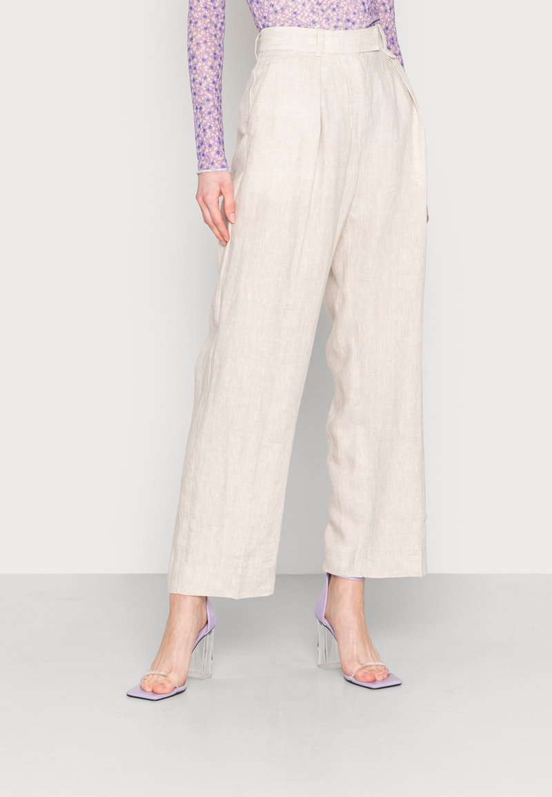 Pantalon large en lin beige avec plis devant, associé à des sandales à talons carré lavande, sur un fond neutre.