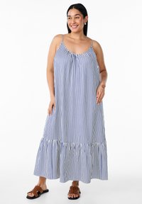 Gestreiftes Sommerkleid in Hellblau und Weiß, mit dünnen Trägern, gerafftem Ausschnitt und einem ausgestellten Rüschensaum. Getragen mit braunen Sandalen.
