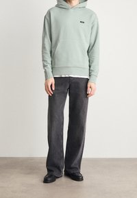 Sudadera verde claro con bolsillo frontal y logo negro, combinada con jeans negros de corte holgado y zapatos negros. Textura suave, estilo casual.