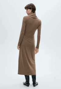 Lang, tætsiddende, brun kjole med høj hals og lange ærmer. Stoffet ser struktureret ud. Båret med sorte knæhøje støvler.