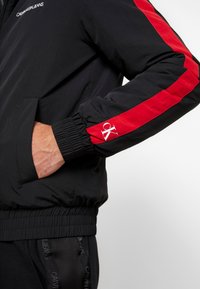 Svart bomberjacka med röda sidostripes, resårsärm och en dragkedjeficka. Har "CALVIN KLEIN JEANS"-logotyp på bröstet och "ck" på ärmen.