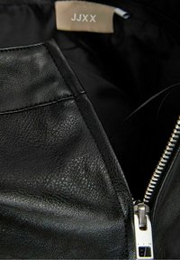 Interior de una chaqueta de cuero negra con una textura suave, un zipper plateado y una etiqueta de tela con "JJXX" en un color claro en la parte superior.