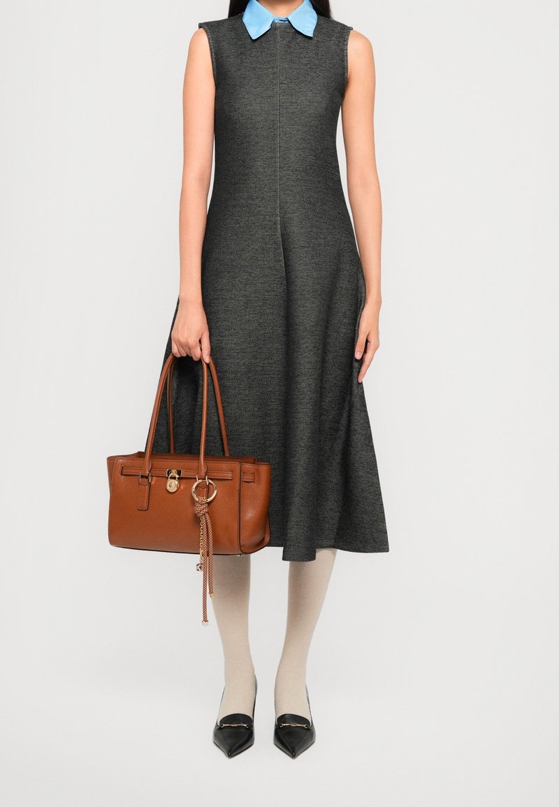 Femme portant une robe sans manches gris foncé avec un col bleu, des collants beiges, des chaussures noires à bouts pointus, et tenant un sac à main en cuir marron.