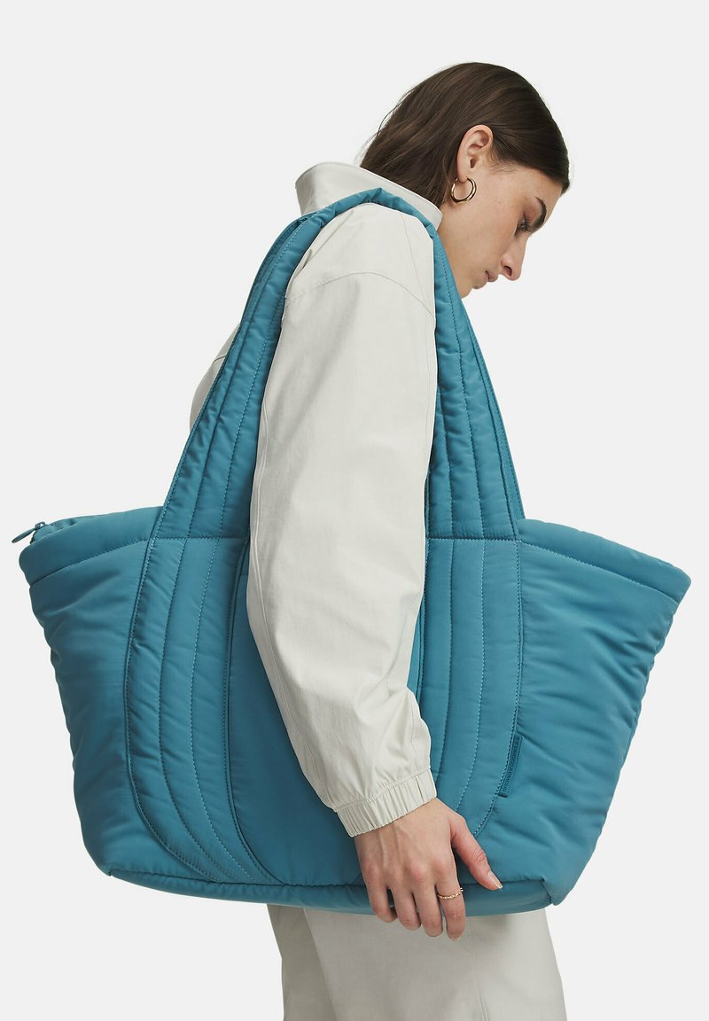 Kvinde iført hvid jakke, bærende en stor, quiltet blå skuldertaske mod hvid baggrund.