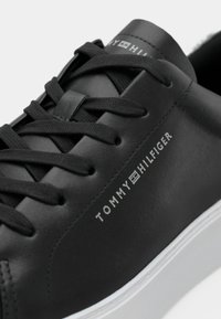 Črne sintetične športne čevlje z ravnimi vezalkami, ki imajo belo gumirano podplat in vtisnjen logotip "TOMMY HILFIGER" na strani. Gladka tekstura.