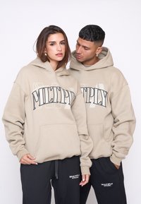 Bežové mikiny s voľným strihom, s veľkým čiernym grafikom "MULTPLY". Materiál sa zdá byť jemný, v kombinácii s čiernymi teplákmi.