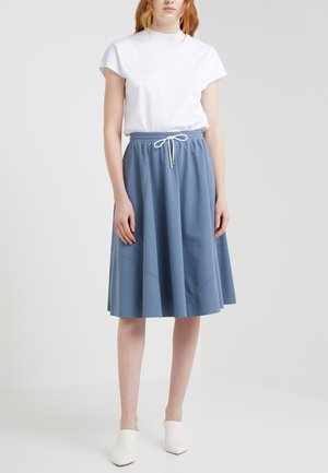 Femme portant un haut blanc à manches courtes, une jupe bleue longueur genou avec une taille à cordon, et des chaussures blanches à bout pointu, debout devant un fond uni.