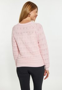 Maglione rosa chiaro con motivi testurizzati orizzontali e verticali, caratterizzato da dettagli a maglia aperta e polsini a coste. Indossato con pantaloni neri.