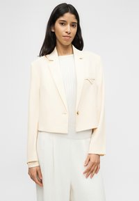 Γυναίκα με μακριά σκούρα μαλλιά, φορώντας ένα κρεμ κοντό σακάκι πάνω από ένα ανοιχτόχρωμο ριμπ πλεκτό τοπ και άσπρο παντελόνι με φαρδιά πόδια, στέκεται μπροστά σε απλό φόντο.