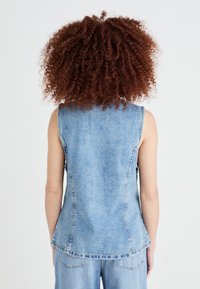 Denim brezrokavnik v svetlo modri barvi, s prilegajočim dizajnom, zadnjimi šivi in ukrivljenim robom, ki se rahlo podaljšuje ob straneh.