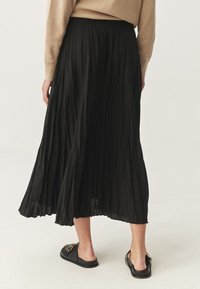 Persona che indossa un maglione beige e una gonna midi plissettata nera in piedi, visibile di spalle, con scarpe slip-on nere con dettagli dorati.