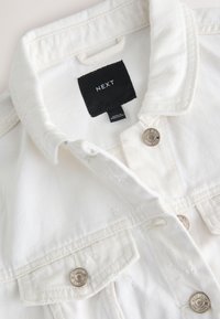 Biała kurtka jeansowa z kołnierzem, z srebrnymi metalowymi guzikami i czarną metką. Tekstura jest gładka, z widocznymi przeszyciami i efektami postarzania.