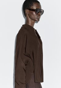 Brązowa, luźna koszula na guziki z wysokim kołnierzem, miękką teksturą i bocznymi rozcięciami; w zestawieniu z dużymi ciemnymi okularami przeciwsłonecznymi.