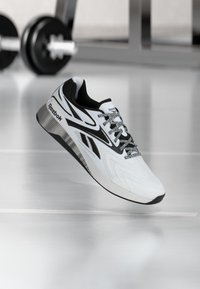 Λευκό και μαύρο αθλητικό παπούτσι Reebok αιωρείται πάνω από το πάτωμα ενός γυμναστηρίου με θολές αλτήρες στο φόντο.