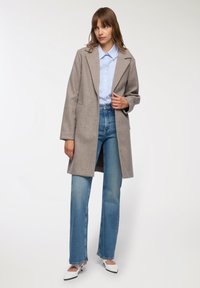 Donna che indossa un cappotto grigio lungo fino al ginocchio sopra una camicia a righe blu, jeans blu dritti e tacchi a punta bianchi su uno sfondo bianco.