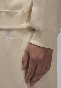 Felpa beige con polsini a costine, caratterizzata da un logo Jumpman ricamato in bianco vicino all'orlo e una texture del tessuto liscia. Mano appoggiata sul tessuto.