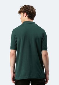 Polo shirt verde scuro realizzato in tessuto liscio, con colletto classico e maniche corte. L'orlo è leggermente più lungo sul retro.