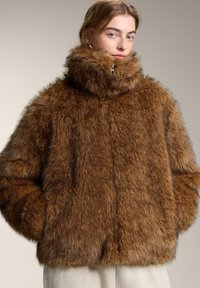 Fiatal személy, aki túlméretezett barna műszőrme kabátot visel, magas gallérral és zsebekbe dugott kézzel, egyszínű háttér előtt.