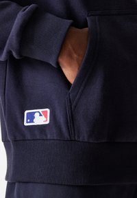 Navy Hoodie aus weichem Stoff mit einer Kängurutasche, gerippten Bündchen und einem weißen MLB-Logo mit roten und blauen Akzenten.