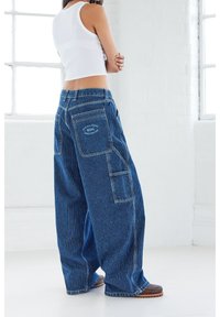 Blå denimöveraller med smala ränder har vida ben och flera fickor. Tyget har en texturerad yta med kontrastsömmar och en logotypmärke.