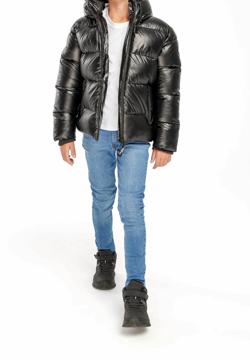 Personne portant une doudoune noire brillante, un t-shirt blanc, un jean skinny bleu et des bottes noires à lacets, debout sur fond blanc.