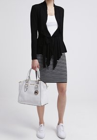 Žena v černém kardiganu přes bílý top, černobílé pruhované sukni, bílých teniskách a držící bílou kabelku se zlatými detaily.