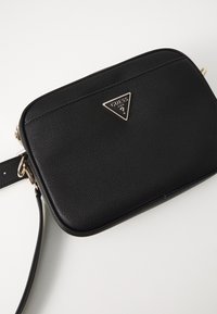 Borsa a tracolla in pelle nera con finitura texturizzata, chiusura con zip e un accento di logo triangolare in oro sul davanti. Include una tracolla regolabile.