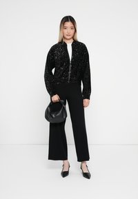Czarna bomberka z cekinami i zamkiem z przodu, zestawiona z dopasowanymi czarnymi spodniami i czarnymi szpilkami. W ręce trzymana jest czarna torebka.