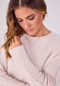 Heller rosa Strickpullover mit gerippten Säumen und strukturiertem Stoff. Das Model hält das Haar und trägt mehrere goldene Halsketten. Weicher, gemütlicher Look.