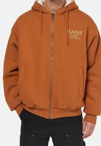 Sudadera con cremallera de forro de felpa marrón, con cordones, dos bolsillos laterales y un logo en el pecho. Presenta una cintura y puños acanalados.