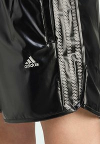 Fekete sportnadrág fényes anyagból, három fehér csíkkal és méhsejt mintával, bal oldalon Adidas logóval.