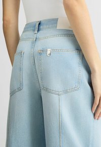 Jeans din denim albastru deschis au talie înaltă, două buzunare pe spate cu clapete pătrate și cusături contrastante, având un eticheta albă la talie.