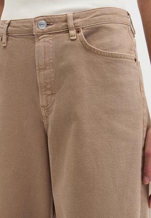 Gros plan sur un jean en denim beige avec bouton de fermeture, poches avant, passants pour ceinture, et une main posée sur le côté.
