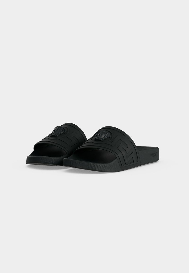 Pair of black slide sandals with a textured strap featuring a raised Medusa head and geometric pattern.