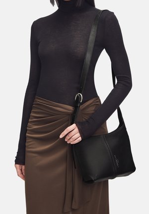 Schwarze Leder-Schultertasche mit einer geschwungenen Form, ausgestattet mit einem langen Riemen und einem dezenten Logo-Detail. Das Modell trägt ein enges schwarzes Oberteil und einen braunen Rock.