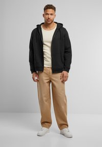 Sort zip-up hættetrøje med lommer, båret over en cremefarvet tanktop, parret med beige bredbenede bukser og hvide sneakers.