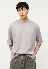 Νεαρός με κοντά σγουρά μαύρα μαλλιά, φοράει μια άνετη ανοιχτό γκρι μπλούζα και ξεθωριασμένο γκρι τζιν, στέκεται με τα χέρια πίσω από την πλάτη του.