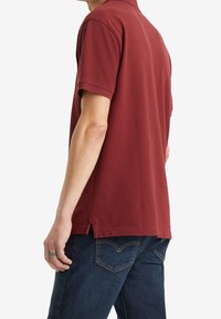 Polo rouge à manches courtes, col côtelé, fentes sur les côtés et finition texturée, associé à un jean bleu foncé avec poches arrondies.