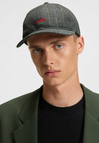 Grey plaid cap with curved brim, featuring red "HUGO" logo. Soft texture, adjustable strap, and a round top with a button detail.