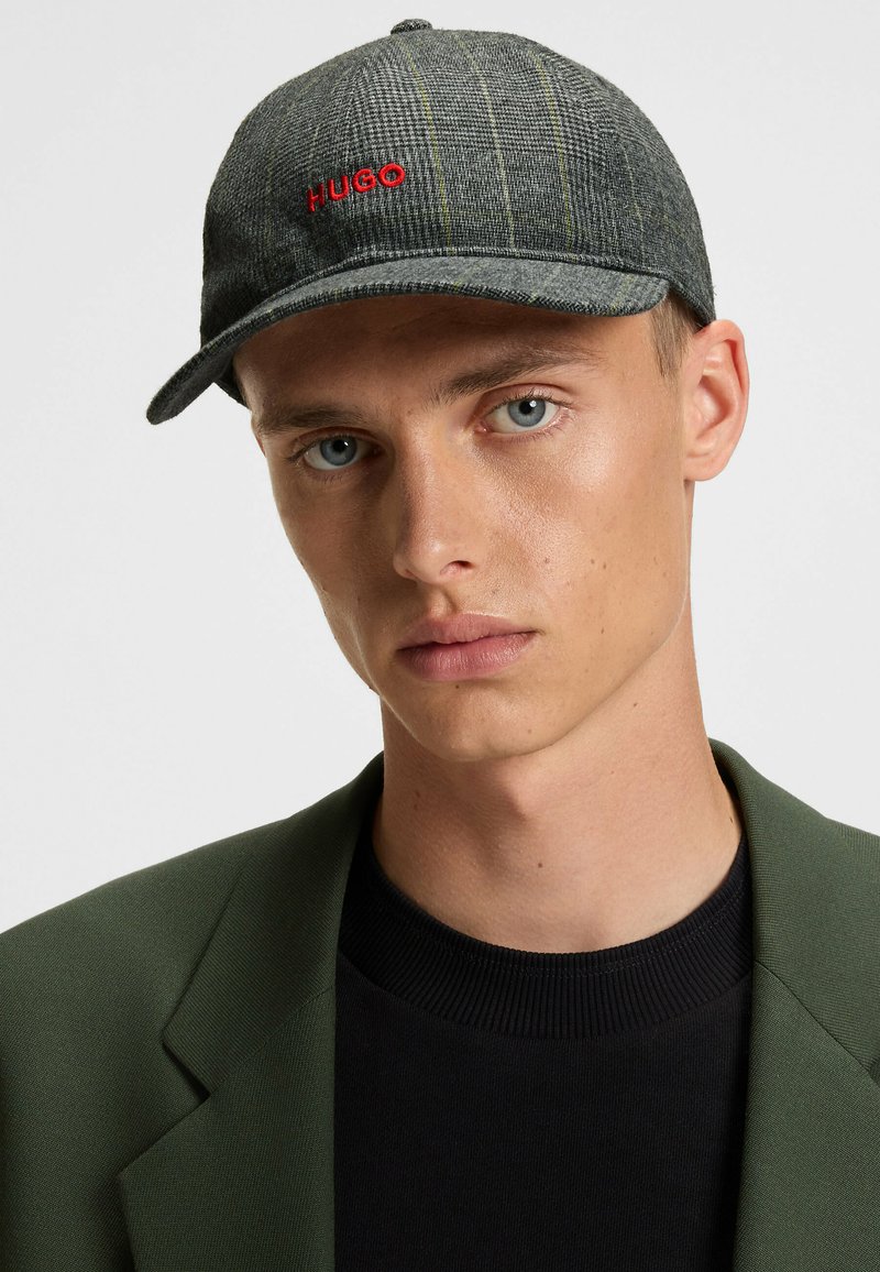 Grey plaid cap with curved brim, featuring red "HUGO" logo. Soft texture, adjustable strap, and a round top with a button detail.