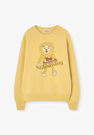 Geel sweatshirt met raglanmouwen heeft een cartoonleeuw-graphic en de tekst "The Animals Observatory" in een speelse lettertype aan de voorkant.