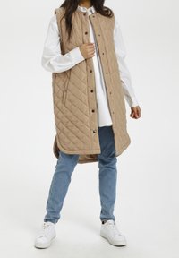 Quilted beige vest with a stand collar, snap buttons, and side pockets, worn over a white shirt and blue jeans, paired with white sneakers.