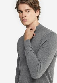 Grauer Strickpullover mit hohem Kragen und langen Ärmeln. Der Stoff wirkt weich mit einer glatten Textur und minimalistischen Details.