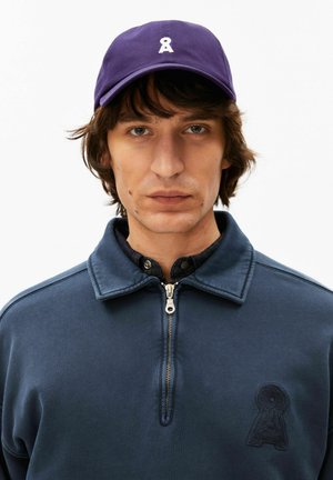 Mann mit mittellangen braunen Haaren, der eine lila Kappe und ein marineblaues Sweatshirt mit Viertel-Reißverschluss und erhabenem Logo auf der Brust trägt.