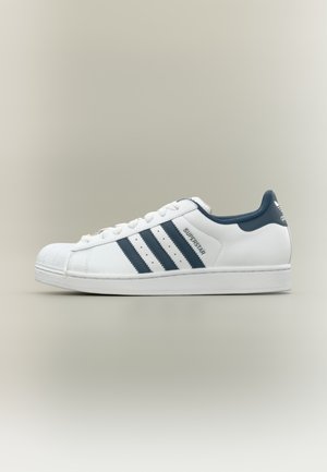 Klassieke witte Adidas Superstar-schoen met drie donkerblauwe strepen, rubberen shell-toe en een bovenwerk van getextureerd leer. Voorzien van een gevoerde kraag en vetersluiting.