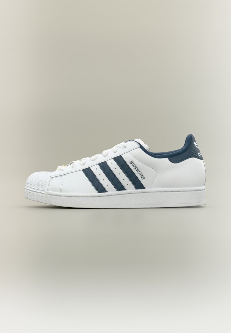 Κλασικό λευκό παπούτσι Adidas Superstar με τρεις σκούρες μπλε ρίγες, λαστιχένια προφυλακτική μύτη και επάνω μέρος από υφή δέρματος. Διαθέτει επενδεδυμένο γιακά και σχέδιο με κορδόνια.